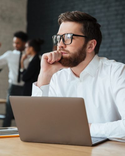 Image of concentrated young caucasian businessman sitting indoors using laptop computer. Looking aside.